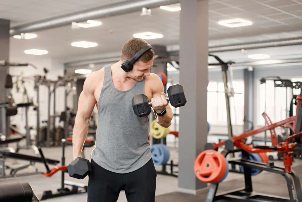 Man in big earphones standing in a modern gym. Man doing excercises with a dumbells. Fit man wearing grey t-shirt.